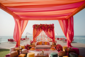 Colorful mehndi tent decor with orange and pink draping, floral canopy, and lounge seating overlooking the ocean at an Indian destination wedding
