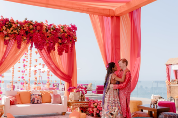 Ummae and Ryan embracing inside a colorful mehndi tent with bright orange and pink draping during an Indian destination wedding celebration
