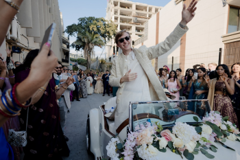 Ryan arriving at his baraat in a vintage white convertible as wedding guests cheer and dance.
