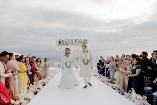 Ummae and Ryan walking down the aisle after their oceanfront wedding ceremony as guests celebrate at their Indian destination wedding at Dreams Bahia Mita Caption: Ummae and Ryan walk hand in hand down the aisle as guests shower them with petals following their oceanfront ceremony.