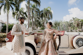 Bride and groom in traditional Indian wedding attire stand by a vintage convertible surrounded by palm trees during a destination wedding photoshoot.