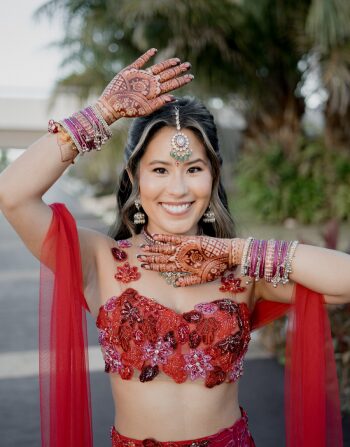Bride in red sangeet outfit posing with detailed mehndi and traditional jewelry during an Indian destination wedding celebration.