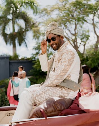 Groom wearing traditional Indian wedding attire and sunglasses arriving for the baraat in a vintage convertible during a destination wedding.