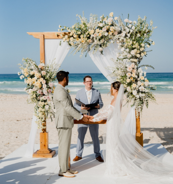 Bride and groom holding hands during a Western wedding ceremony under a floral arch by the ocean at a destination wedding.
