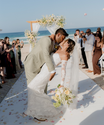 Bride and groom sharing a dip kiss during a beachside wedding ceremony aisle moment with guests celebrating around them