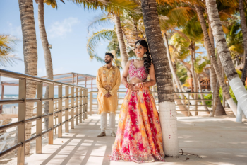 Indian couple posing by palm trees in colorful haldi outfits at a beach resort before wedding festivities.