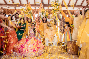Indian haldi ceremony with bride and groom surrounded by family throwing yellow flower petals in a joyful celebration
