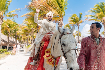 Indian groom riding white horse during baraat procession with guests dancing at destination wedding.