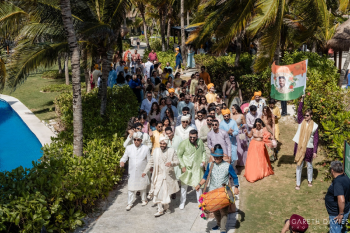 Large Indian wedding baraat procession with guests dancing, drummer leading, and custom flag with groom’s image.