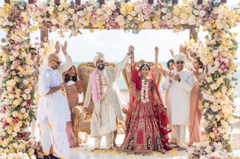 Bride and groom at mandap raising hands in celebration after Indian wedding ceremony with floral mandap and ocean backdrop