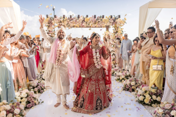 Indian bride and groom smiling and walking down aisle after beachfront wedding ceremony surrounded by guests and floral décor at El Dorado Royale resort.
