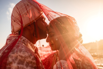 Indian bride and groom in red bridal lehenga and ivory sherwani sharing intimate forehead kiss under bridal dupatta with sunset light during Indian destination wedding in Mexico.