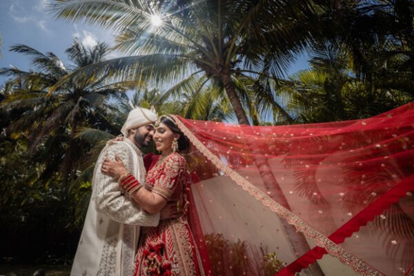 Indian bride and groom in red bridal lehenga and ivory sherwani embracing under palm trees with flowing red dupatta during a destination wedding at El Dorado Royale resort in Mexico.