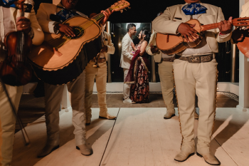 Indian couple sharing a romantic moment encircled by a mariachi band in white suits during a rooftop welcome party in Mexico, bride in burgundy lehenga and groom in cream suit