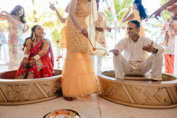Indian bride in red saree and groom in white kurta seated in ornate gold basins laughing as family members apply turmeric paste during an outdoor haldi ceremony at Palapa Ganesha at Generations Riviera Maya surrounded by palm trees.