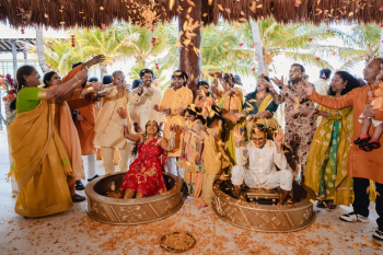 Large group of wedding guests in yellow and orange throwing flower petals over a laughing Indian bride and groom seated in golden haldi tubs during an outdoor ceremony with tropical palm trees