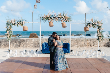 Indian bride in a teal embroidered lehenga and groom in a navy bandhgala jacket posing on a wooden dance floor at a beachside sangeet setup with blue velvet sofas, floral arches, and the Caribbean sea behind them