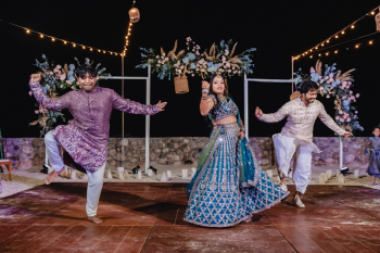 Indian bride in a teal and silver embroidered lehenga dancing energetically at an outdoor sangeet night at El Dorado Royale with guests, string lights overhead and floral decorations visible in the background.
