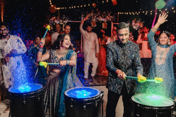Indian bride and groom laughing and playing illuminated LED water drums at a sangeet celebration at night, drums glowing blue and green as water splashes around them
