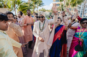 Indian groom in cream sherwani and sunglasses dancing and celebrating with groomsmen and guests during a baraat procession at Generations Riviera Maya, palm trees and a resort building visible in the background