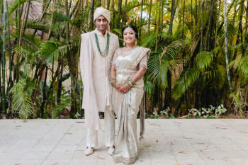 Indian bride and groom standing together in a formal portrait among bamboo and tropical greenery, groom in a cream embroidered sherwani with green gemstone mala and bride in a silver-grey ceremonial saree with gold embroidery