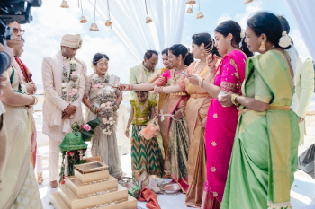 Hindu wedding ceremony ritual on the beach under a white draped mandap with golden bells, Indian bride and groom surrounded by family members performing traditional rites, Caribbean ocean visible in the background