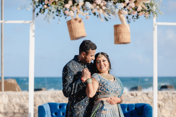 Indian bride and groom pose together before their sangeet, with ocean views behind them and floral décor overhead.