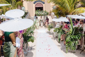 Indian bride in a white and silver embroidered wedding lehenga walking down a white sand aisle flanked by guests holding white parasols and tall tropical greenery arrangements, with rose petals scattered along the path