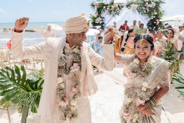 Sanjana and Hirsh celebrating after their beach wedding ceremony at Generations Riviera Maya, wearing matching white and cream embroidered sherwani and lehenga, with the Caribbean ocean and floral mandap visible behind them.