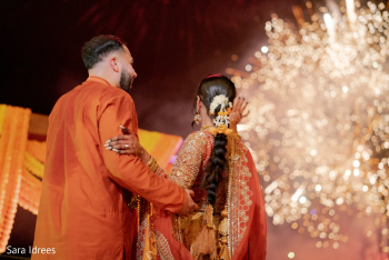 Aninder and Navneet playing drums during a lively sangeet or reception celebration, surrounded by dancing guests under string lights and colorful powder effects.
