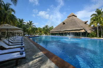 Tropical pool area at El Dorado Royale with lounge chairs, palm trees, and a large palapa-style building creating a relaxing atmosphere for destination wedding guests. Tropical pool area at El Dorado Royale with lounge chairs, palm trees, and a large palapa-style building creating a relaxing atmosphere for destination wedding guests.