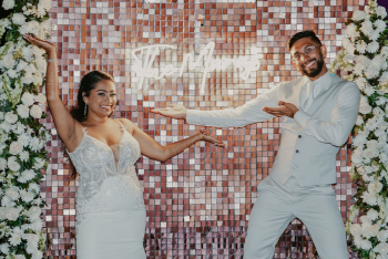 Malena and Eshwar posing in front of their reception photo wall framed by flowers during their rooftop wedding reception