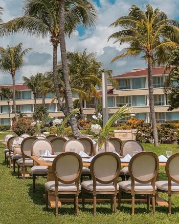 Outdoor garden venue setup with long banquet tables, floral centerpieces, and palm trees creating an elegant event space