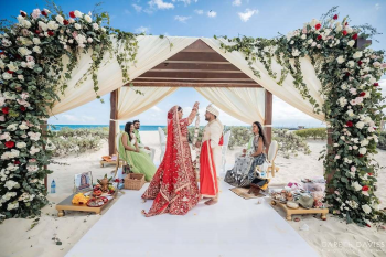 Bride and groom exchanging a floral jaimala under a beachfront floral mandap at Dreams Playa Mujeres, with family members seated on either side during the South Asian wedding ceremony. Bride and groom exchanging a floral jaimala under a beachfront floral mandap at Dreams Playa Mujeres, with family members seated on either side during the South Asian wedding ceremony.