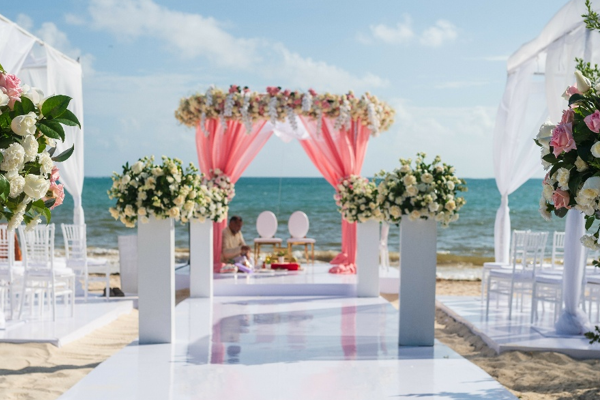 Oceanfront mandap decorated with coral drapery and floral pillars at a South Asian wedding ceremony on the beach at Royalton Splash Riviera Cancun, Mexico.