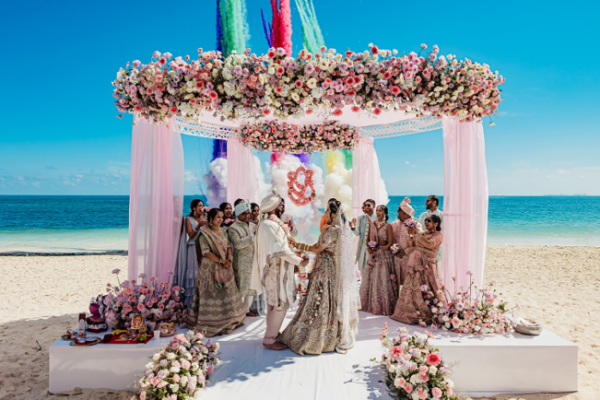 Roshni and Paraj exchange vows beneath a floral mandap at Planet Hollywood Cancun during their Indian destination wedding in Mexico, with colorful smoke effects creating a dramatic backdrop overlooking the Caribbean.
