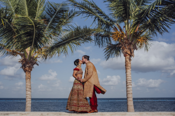 Seema and Logan kissing between palm trees in traditional Indian wedding attire with the Caribbean Sea behind them at Moon Palace Cancun. Seema and Logan kissing between palm trees in traditional Indian wedding attire with the Caribbean Sea behind them at Moon Palace Cancun.