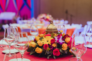 Table setting inside a ballroom for a Sangeet with red table linens and a centerpiece with red, yellow, and purple orchids and a golden lantern. Table setting inside a ballroom for a Sangeet with red table linens and a centerpiece with red, yellow, and purple orchids and a golden lantern.