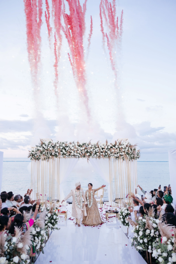 Beachfront gold mandap with pampas grass with pink smoke bombs and sparklers going off behind it for an oceanfront Indian wedding ceremony Beachfront gold mandap with pampas grass with pink smoke bombs and sparklers going off behind it for an oceanfront Indian wedding ceremony