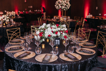 Table settings at an Indian wedding reception with black linens, gold charger plates, a “Thank You” note to guests and a centerpiece with red and white flowers. Table settings at an Indian wedding reception with black linens, gold charger plates, a “Thank You” note to guests and a centerpiece with red and white flowers.