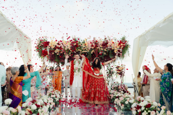 Bhavya and Leon wave to their cheering guests as rose petals rain down after their South Asian wedding ceremony at AVA Resort Cancun on the Mexican Caribbean, with a suspended red and blush floral mandap and a mirrored aisle. Bhavya and Leon wave to their cheering guests as rose petals rain down after their South Asian wedding ceremony at AVA Resort Cancun on the Mexican Caribbean, with a suspended red and blush floral mandap and a mirrored aisle.