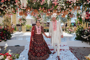 Smiling bride in a maroon lehenga and groom in an ivory sherwani hold hands on a rose petal-covered platform under a gold mandap draped with pink and white florals and rattan lanterns at their Indian wedding ceremony at Hard Rock Hotel Los Cabos, Mexico. Smiling bride in a maroon lehenga and groom in an ivory sherwani hold hands on a rose petal-covered platform under a gold mandap draped with pink and white florals and rattan lanterns at their Indian wedding ceremony at Hard Rock Hotel Los Cabos, Mexico.